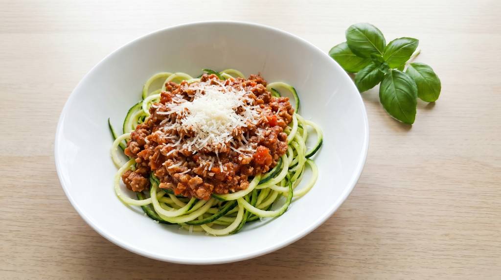 Truthahn-Bolognese mit Zucchini-Nudeln und geriebenem Parmesan in einer weißen tiefen Schüssel, Draufsicht auf hellem Holzuntergrund